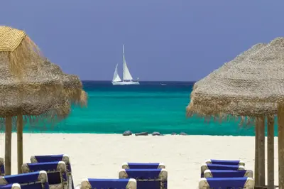 a beach with chairs and umbrellas and a sail boat in the water at Agua Hotels Sal Vila Verde in Santa Maria