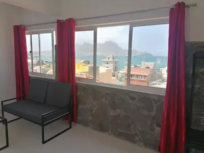 a chair in front of a window with red curtains at STUDIO VUE MER - CASA da LUZ in Mindelo