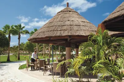 a man sitting at a table at a restaurant with a thatch roof at Hotel Riu Cabo Verde - Adults Only - All Inclusive in Santa Maria